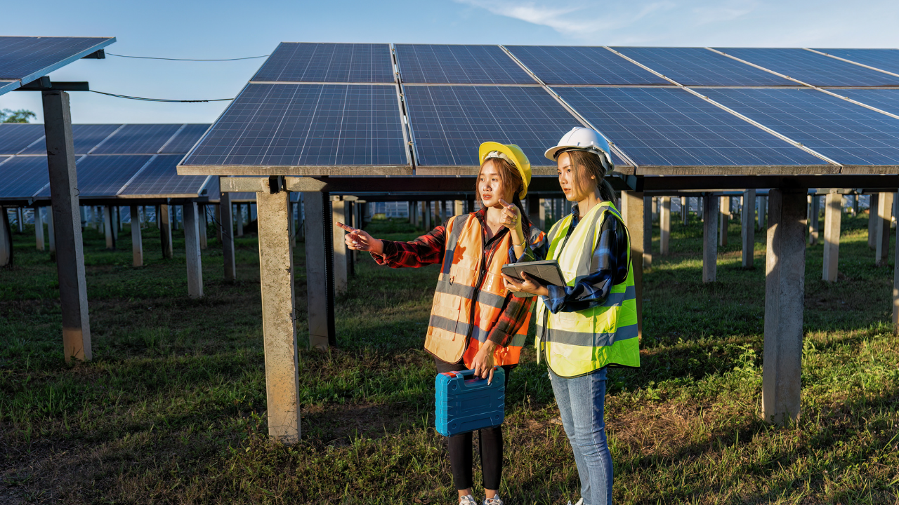 Dos mujeres con chalecos reflectantes y cascos se comunican mientras observan una instalación de paneles solares. La escena refleja una jornada laboral en terreno, con atención al detalle y actitud resolutiva, característica del enfoque español.