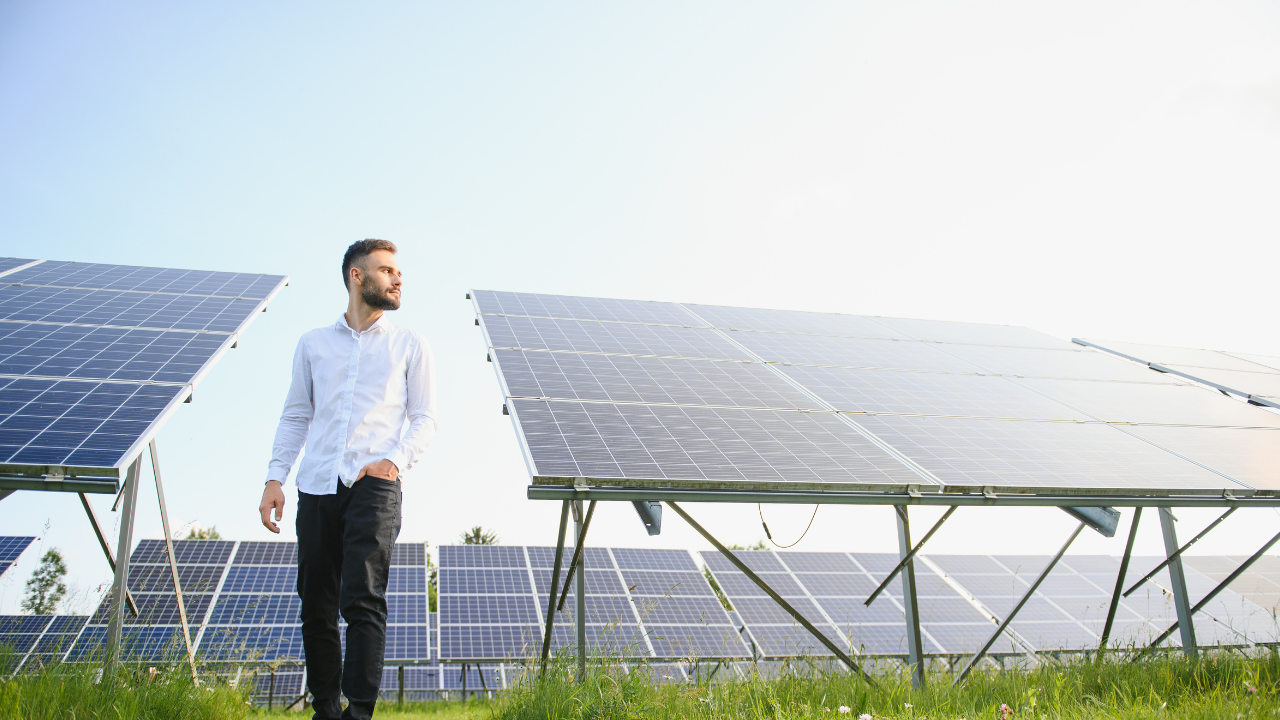 A man in a white shirt walks confidently between solar panels under a clear sky. The image portrays an independent solar panel installer surveying his installation site, reflecting American entrepreneurial spirit.