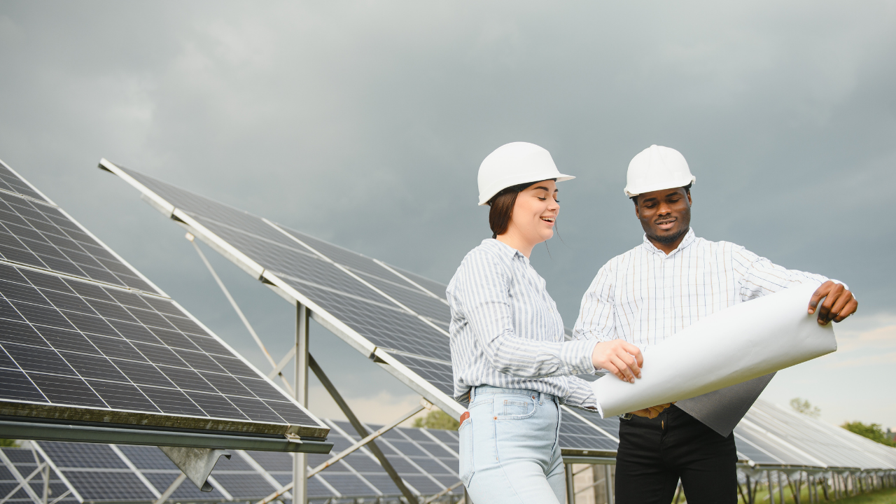 Un hombre y una mujer revisan planos junto a una gran estructura de paneles solares. Su expresión y lenguaje corporal transmiten entusiasmo por los avances en energía renovable, destacando la participación activa de Uruguay en esta transformación.