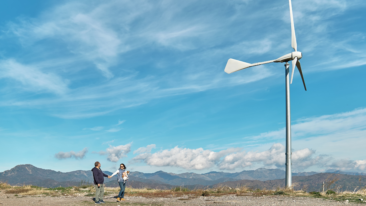 Una pareja conversa cerca de una turbina eólica en un entorno montañoso, evocando el impacto comunitario del desarrollo energético sostenible.
