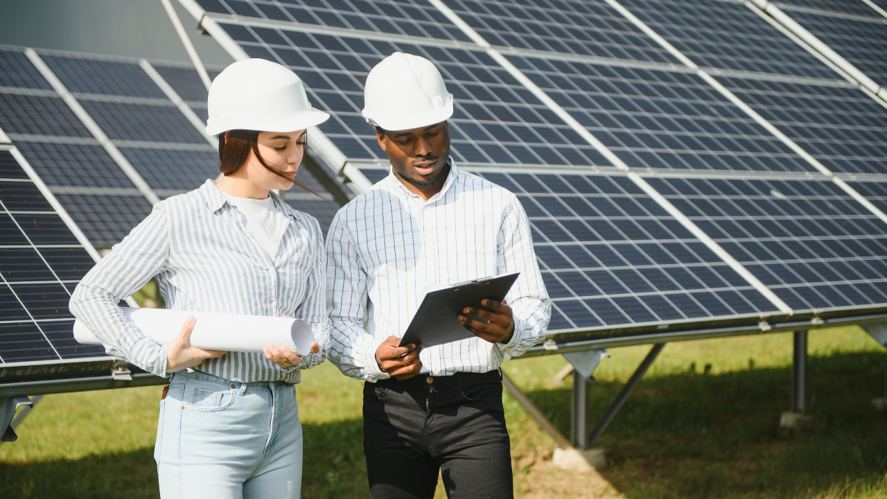 Dos técnicos con cascos blancos revisan documentos frente a una serie de paneles solares. La imagen muestra el trabajo conjunto en un servicio de instalación solar, en un entorno profesional y soleado.