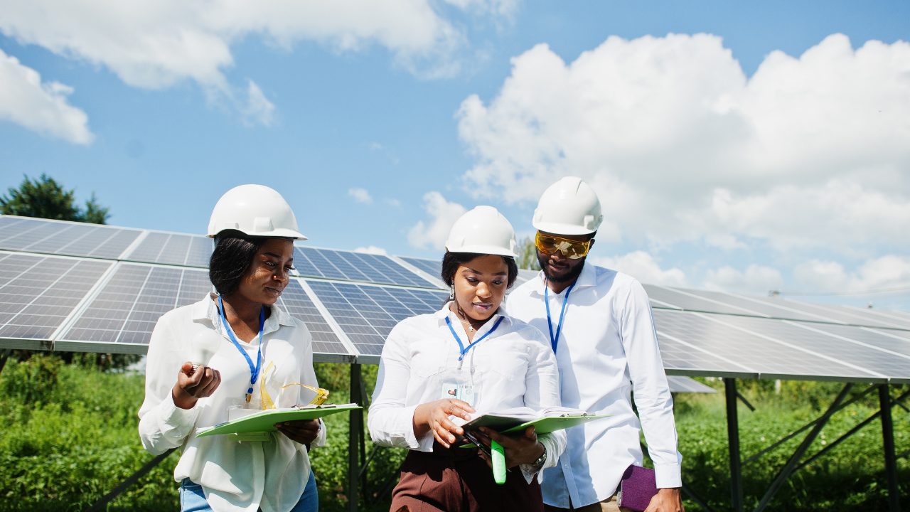 Tres personas con credenciales y cascos revisan documentos en un campo solar. La escena muestra un equipo diverso de instalador de paneles solares trabajando con compromiso bajo el cielo uruguayo.