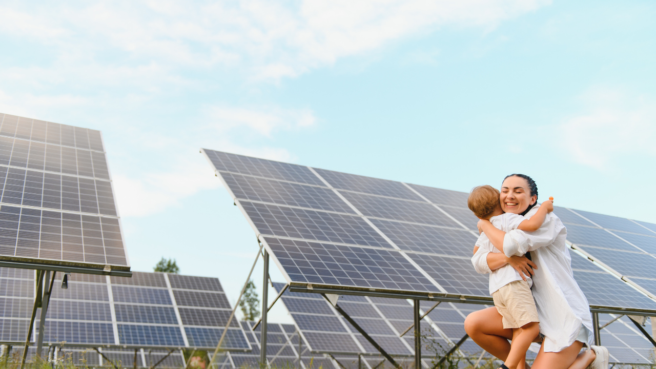 A smiling woman lifts a child near a field of solar panels, symbolizing a clean energy future and family well-being in a bright British landscape.