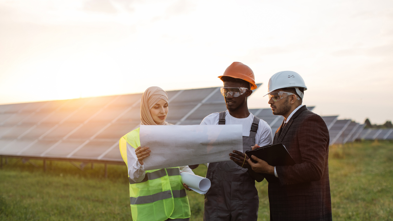 Three professionals from diverse backgrounds analyze blueprints in front of solar panels at sunset, emphasizing strategic efforts to reduce the Carbon footprint through renewable energy collaboration.