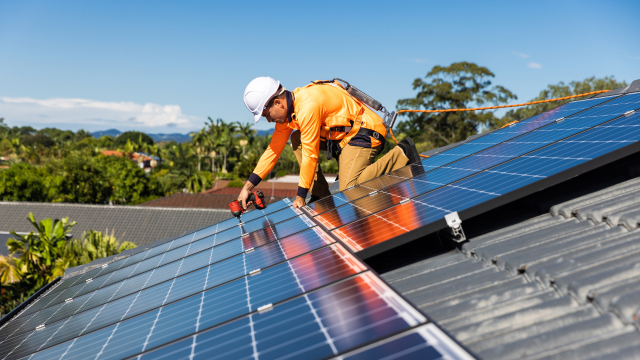 Un instalador en uniforme naranja trabaja sobre un techo metálico rodeado de naturaleza, asegurando los paneles solares usados. Esta imagen representa la apuesta uruguaya por la energía renovable doméstica, integrando tecnología reutilizada en armonía con el entorno.