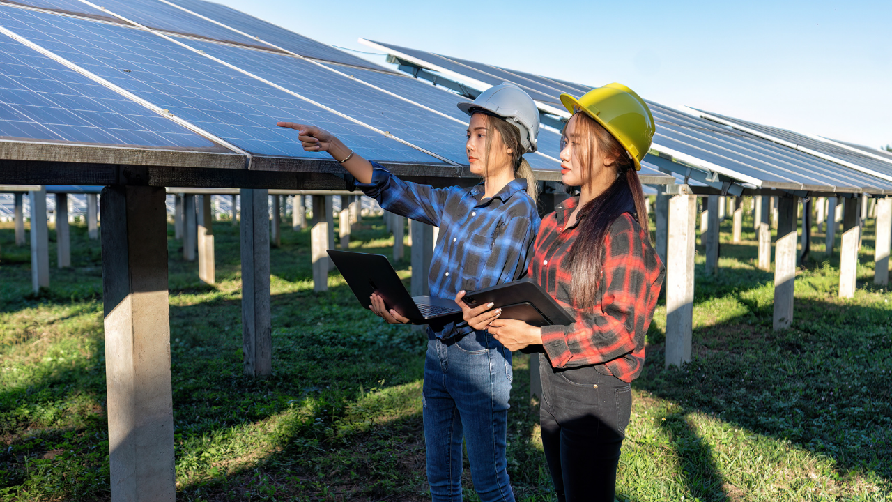 Dos mujeres con portátiles y cascos inspeccionan una planta solar, reflejando el liderazgo técnico femenino en proyectos que buscan mitigar la huella de carbono en Uruguay.