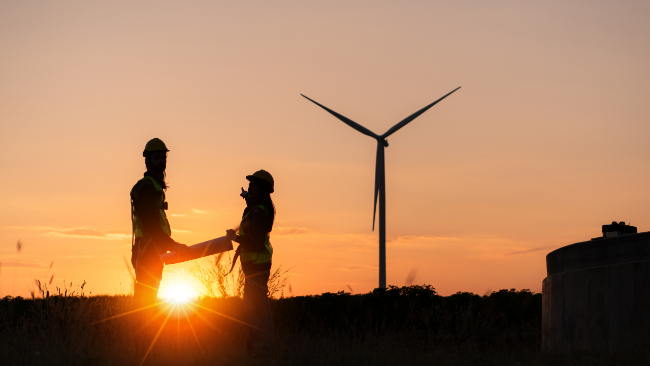 Silhouetted against a sunset, two workers review plans near a wind turbine, capturing the U.S. commitment to wind integration at all stages of the day.