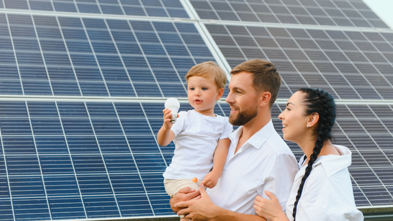 Un niño sostiene una bombilla mientras es cargado por su padre, acompañado de su madre, frente a paneles solares. La imagen transmite la conexión familiar con el futuro energético y el valor del autoconsumo solar en la vida cotidiana.