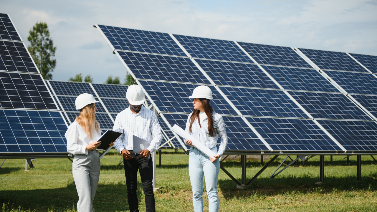 A group of three engineers stands in front of large solar panels, examining blueprints and tablets under a partly cloudy sky, symbolizing teamwork and technological focus on solar panels.