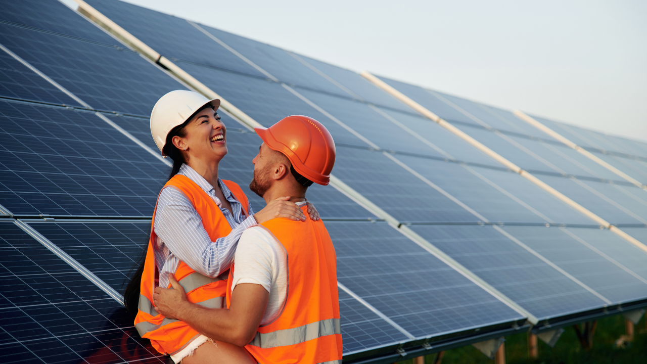Two workers in orange safety vests and helmets share a joyful moment in front of rows of solar panels, blending industrial progress with human connection.