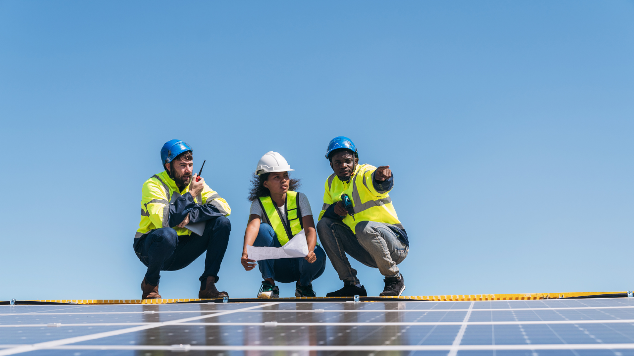 Tres ingenieros se encuentran agachados sobre una instalación de solar panels, analizando planos y conversando con atención, reflejando el trabajo coordinado en el desarrollo de energías renovables.