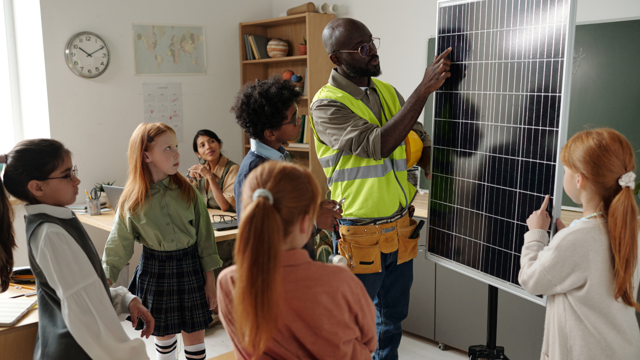 Un operario con chaleco reflectante enseña un panel solar a un grupo de niños en un aula, promoviendo la conciencia sobre paneles solares desde la educación.