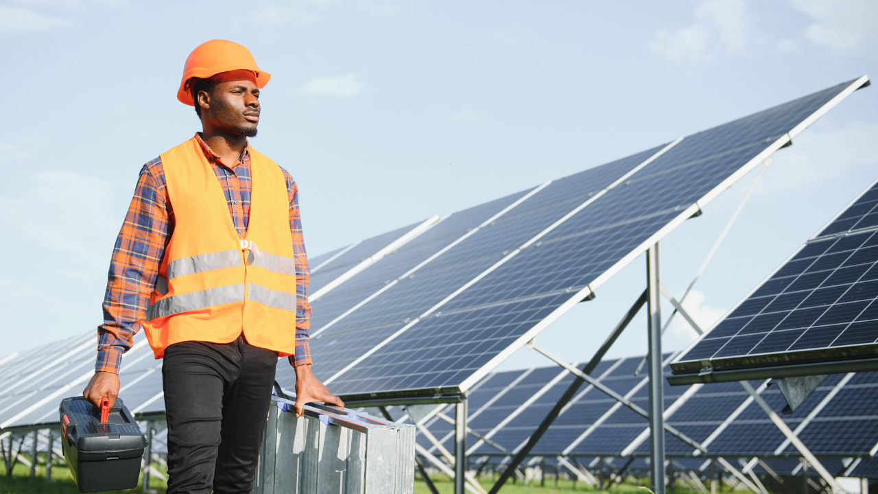A technician in an orange vest and helmet walks confidently between rows of solar panels, carrying tools and materials. The scene highlights practical implementation, symbolizing investment and commitment to finance solar installation on a larger scale.