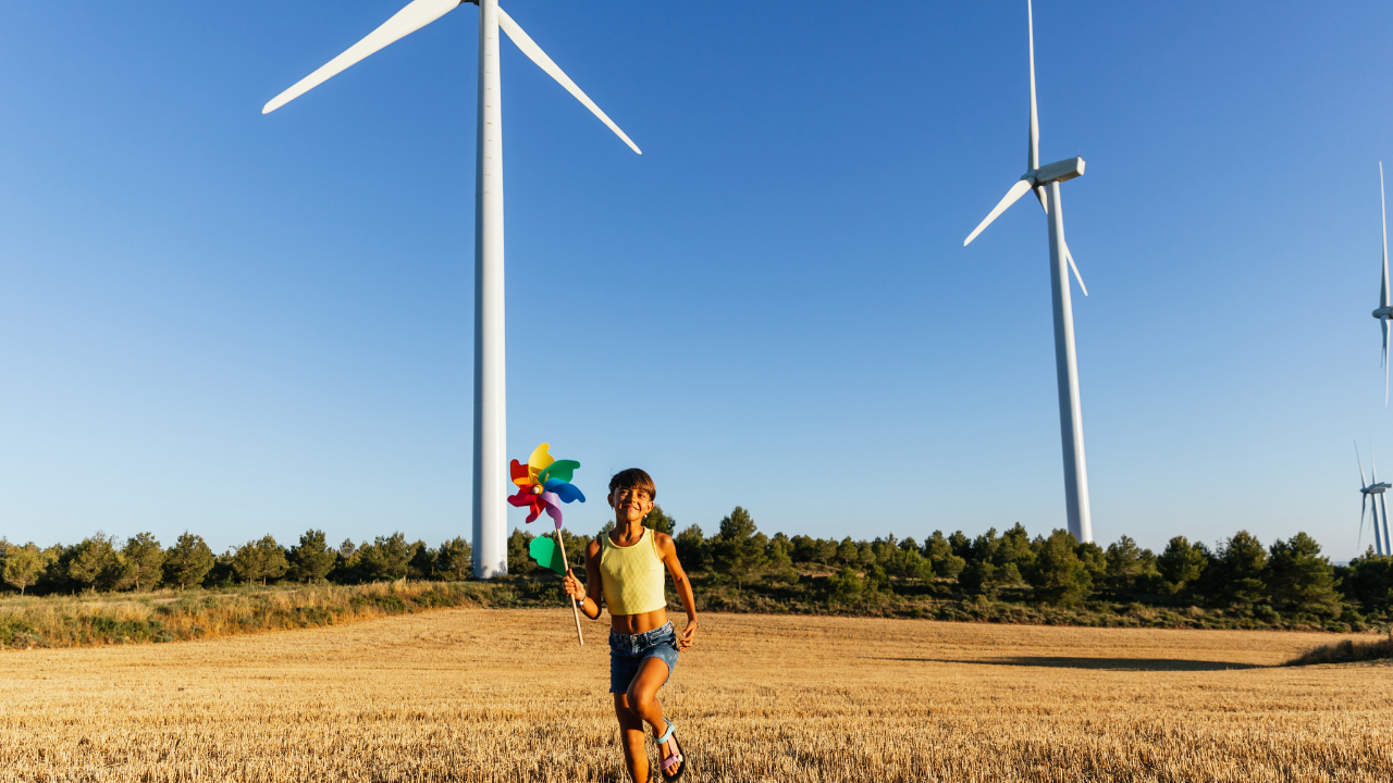 Una niña corre con un molinillo de viento frente a turbinas reales, simbolizando el futuro sostenible que garantiza el mantenimiento de aerogeneradores.
