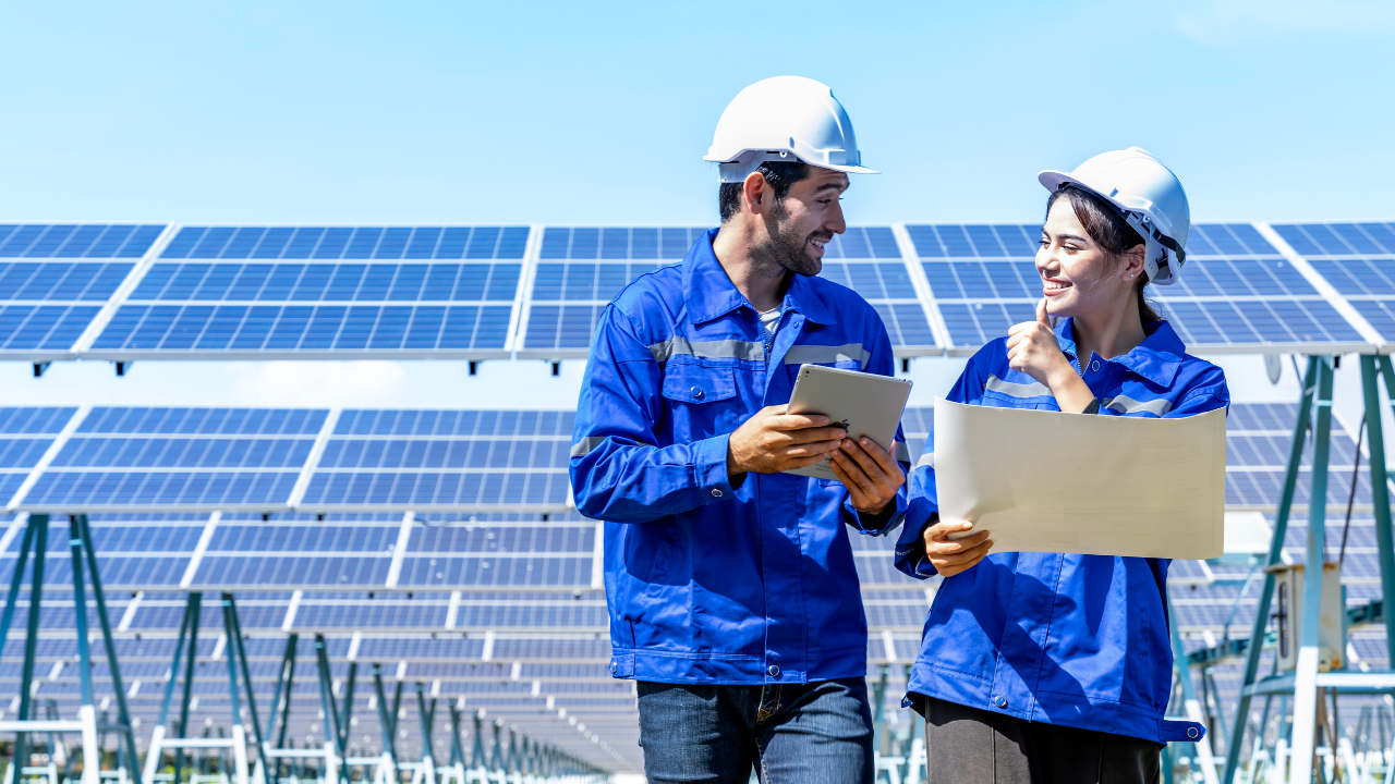 Dos ingenieros sonríen mientras consultan planos y una tablet frente a múltiples paneles solares. La imagen comunica colaboración y planificación, reflejando cómo financiar la instalación solar se traduce en progreso tecnológico y energético en el país.