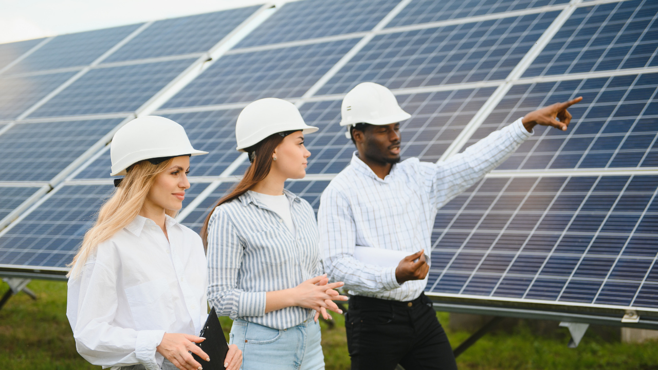 Tres personas con cascos blancos observan atentamente un parque solar; uno de ellos señala hacia adelante. La escena sugiere análisis y visión a futuro, transmitiendo la importancia de financiar la instalación solar para alcanzar objetivos sostenibles.