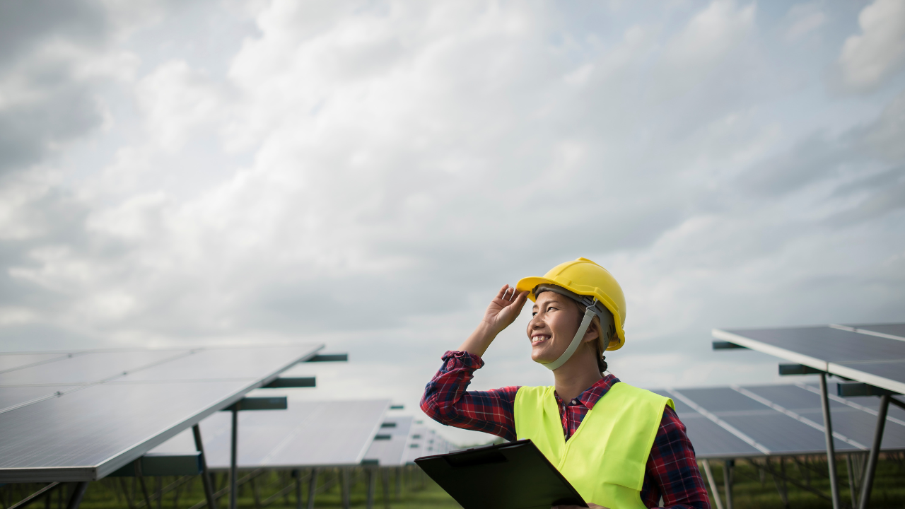 Una joven profesional sonríe en un parque solar, con casco y portapapeles en mano, reflejando optimismo y profesionalismo en torno al desarrollo del autoconsumo solar en Uruguay bajo un cielo parcialmente nublado.