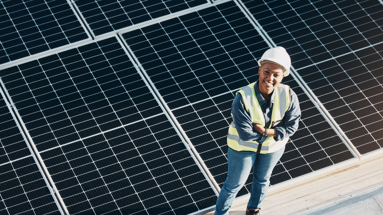 Una mujer sonriente, con casco y chaleco reflectante, posa frente a una matriz de paneles solares. Su expresión transmite confianza y optimismo en el papel de la energía solar en empresas dentro del modelo energético uruguayo.