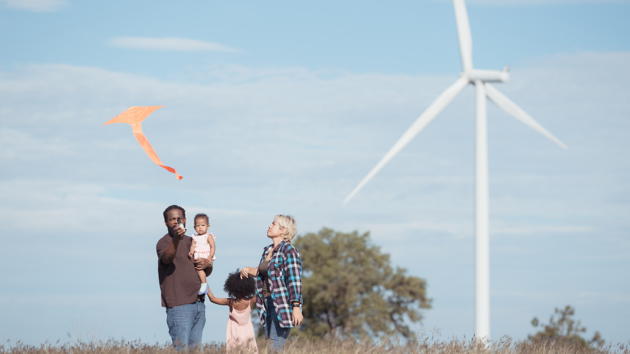 Una familia juega con una cometa en un campo abierto mientras un aerogenerador se alza al fondo, simbolizando la convivencia entre energía renovable y vida cotidiana.