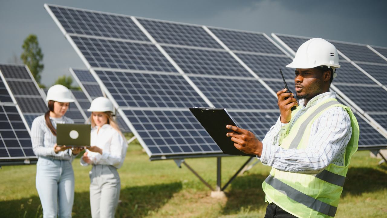 An engineer in a safety vest and white helmet checks a clipboard while speaking into a radio. Behind him, two women in hard hats work with a laptop near solar panels, portraying a structured and professional solar panel installation environment.