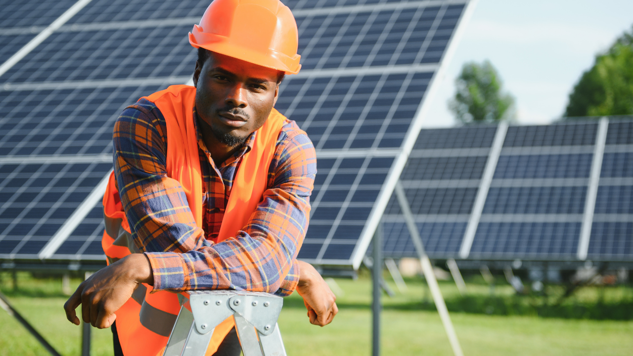 Un trabajador con chaleco naranja y casco posa pensativo frente a paneles solares. La imagen transmite orgullo y compromiso con la instalación de paneles solares, en un entorno rural que sugiere desarrollo sustentable.