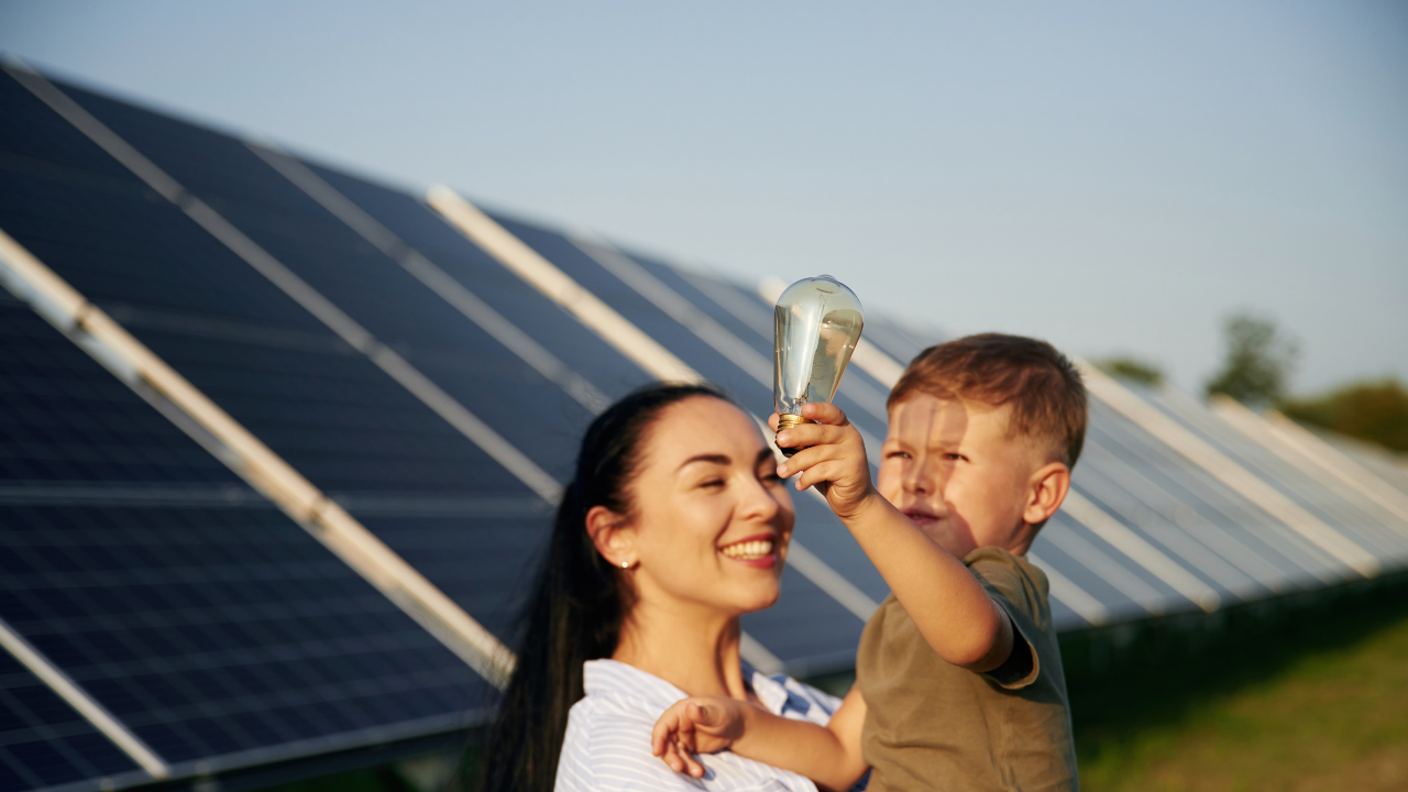 A smiling mother holds her child as he lifts a lightbulb in front of solar panels, representing the idea of excess solar panel energy being passed on to future generations in a hopeful, educational context.