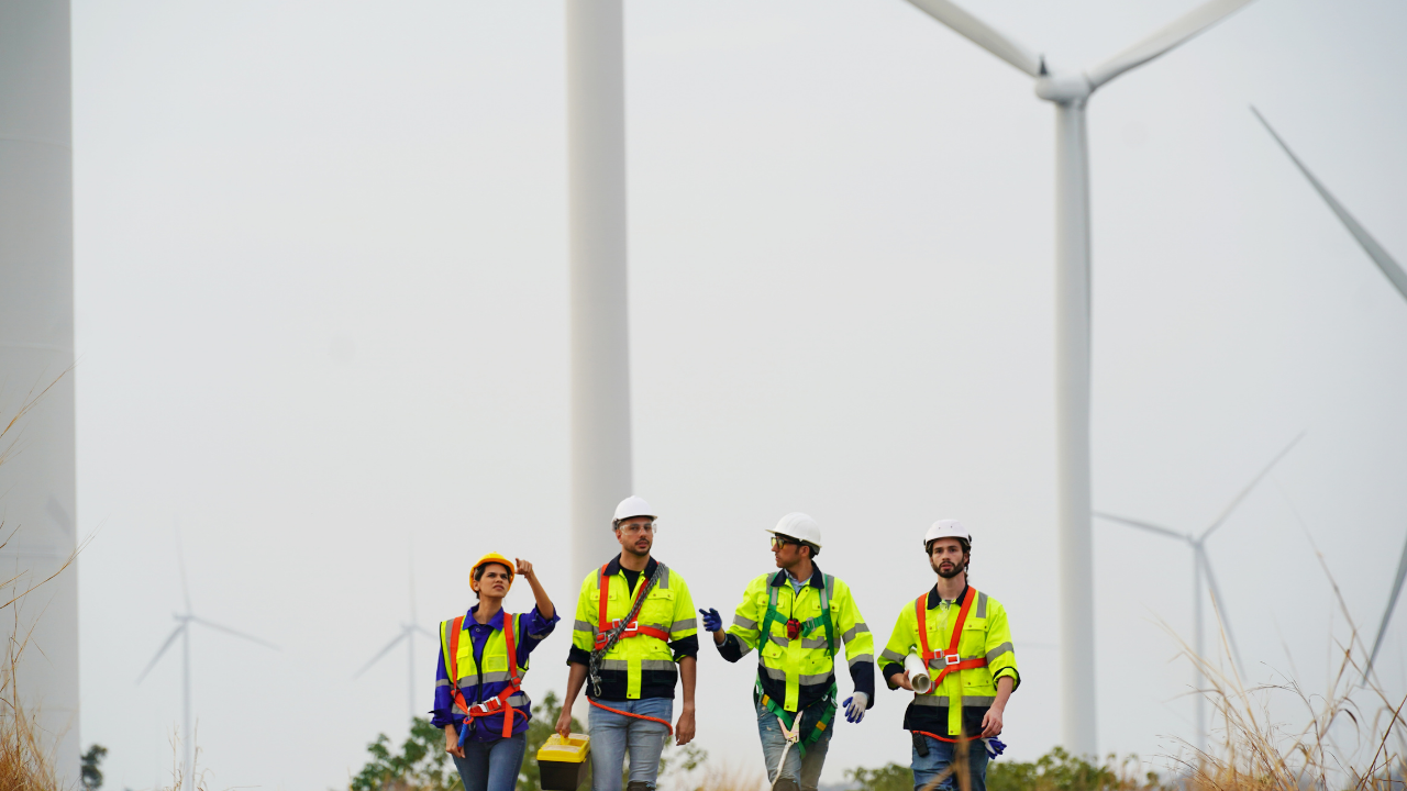 Four wind energy workers walk confidently through a wind farm, reinforcing the importance of home wind energy in the workforce and green technology sector.