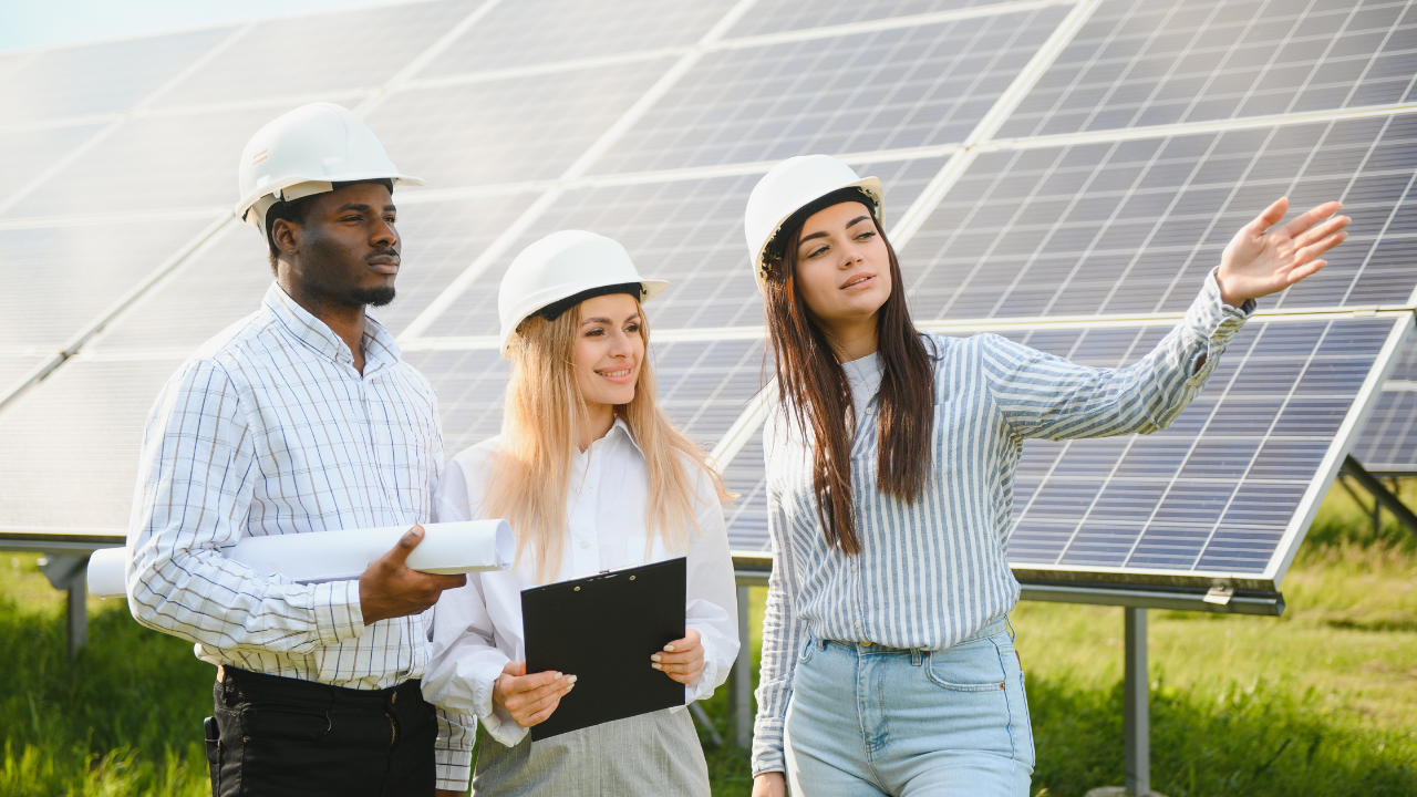 Three engineers wearing hard hats stand beside solar panels, with one pointing ahead. Their attentive expressions suggest a collaborative effort to harness and distribute excess solar panel energy through strategic planning.