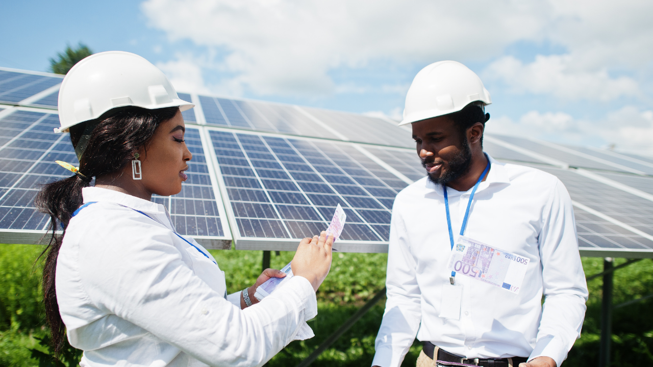 Dos personas con cascos y planos conversan frente a paneles solares, mostrando billetes de euro. La escena representa cómo la energía sobrante paneles solares puede traducirse en valor económico y oportunidades de inversión.