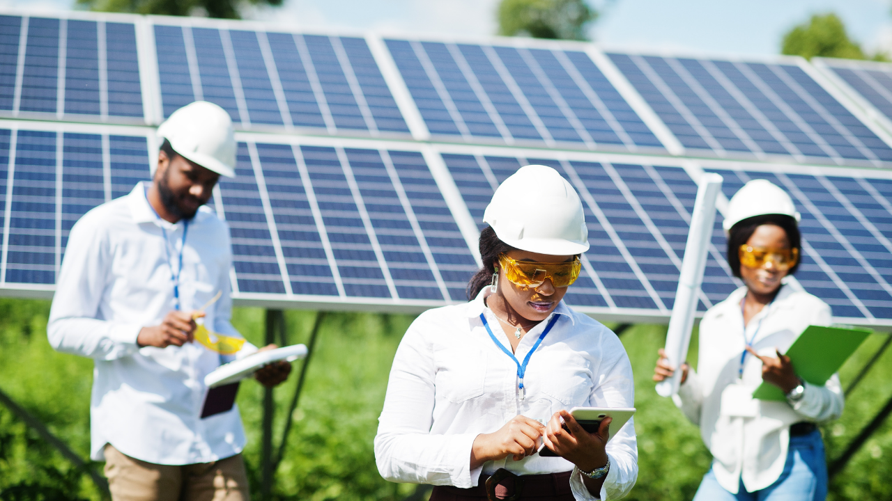 Three engineers, equipped with hard hats and safety goggles, inspect solar shingles in a sunny field. One uses a tablet, another takes notes, and a third holds a document tube, all focused on evaluating renewable energy installations.