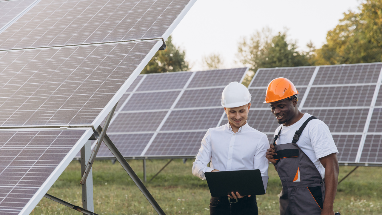Two men stand by large solar panels, discussing project details on a laptop. Both wear hard hats—one in formal attire and the other in work overalls—symbolizing collaboration in implementing solar shingles technology.