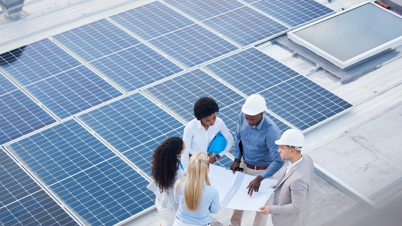 Un grupo de profesionales analiza planos sobre un tejado cubierto de tejas solares. Con cascos de seguridad y documentos en mano, debaten posibles ajustes técnicos en una reunión técnica al aire libre.