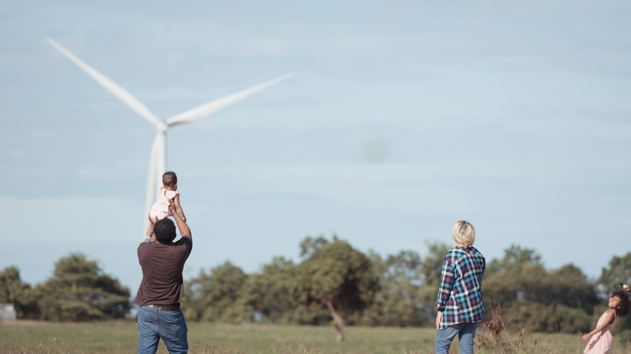 A family enjoys time outdoors under a clear sky, with wind turbines in the background representing the promise of wind energy.