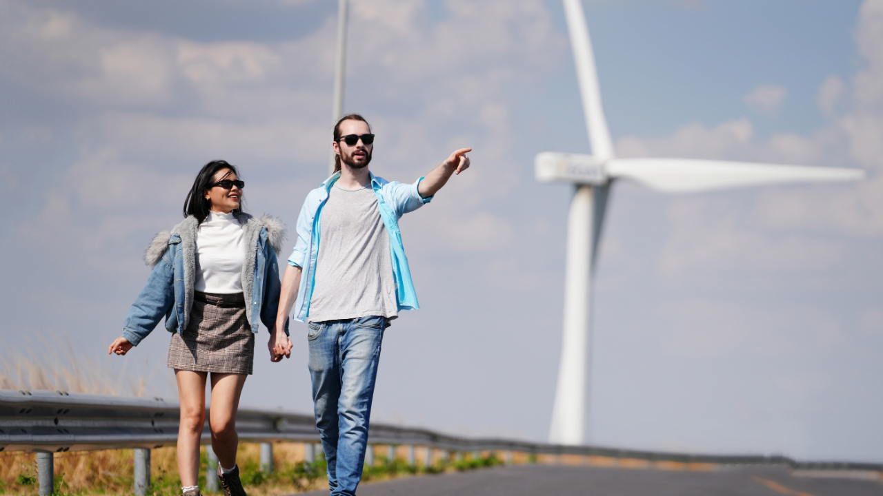 Una pareja camina junto a una carretera rural con aerogeneradores al fondo, integrando la energía eólica en su entorno cotidiano.