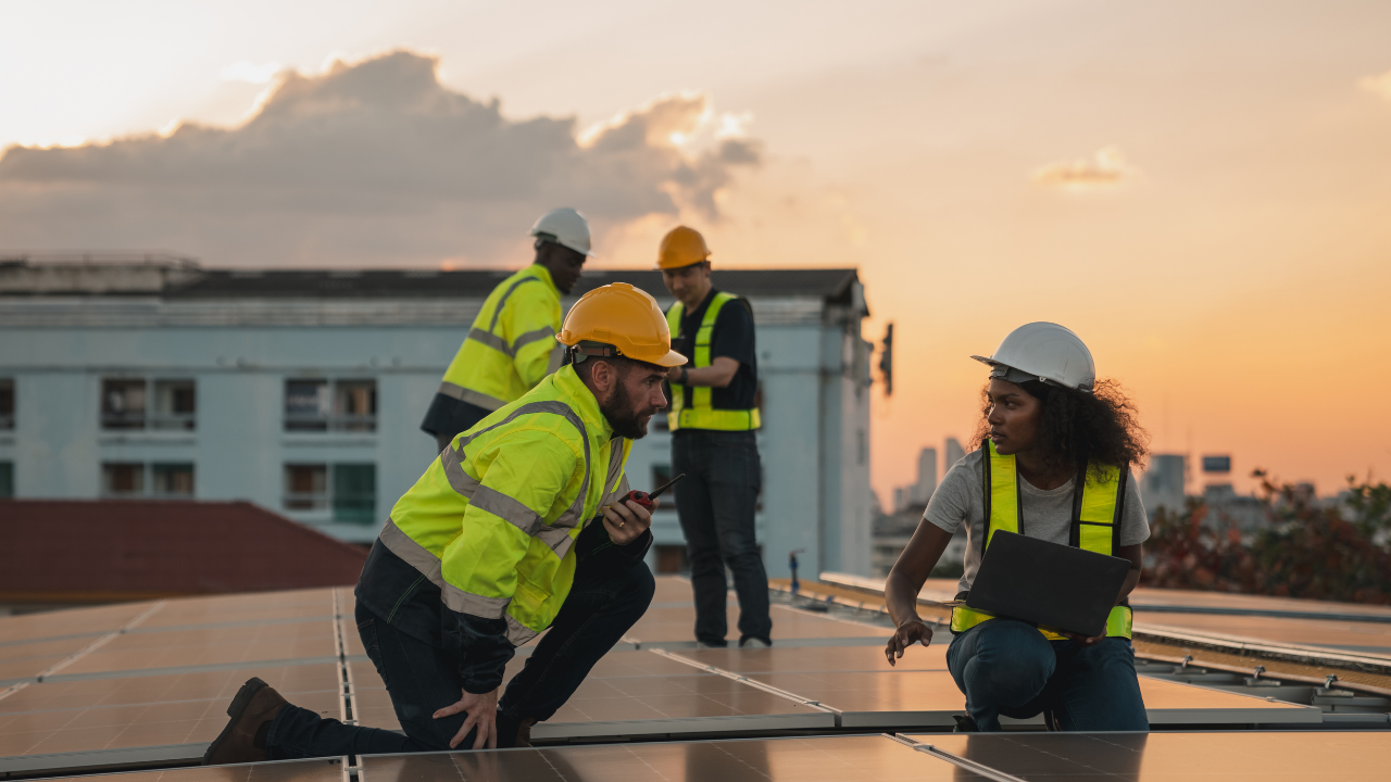 Trabajadores con cascos y chalecos reflectivos dialogan sobre un techo al atardecer, rodeados de paneles solares. La frase "limpiar paneles solares" enfatiza el trabajo colaborativo y el compromiso con la energía limpia en entornos urbanos.