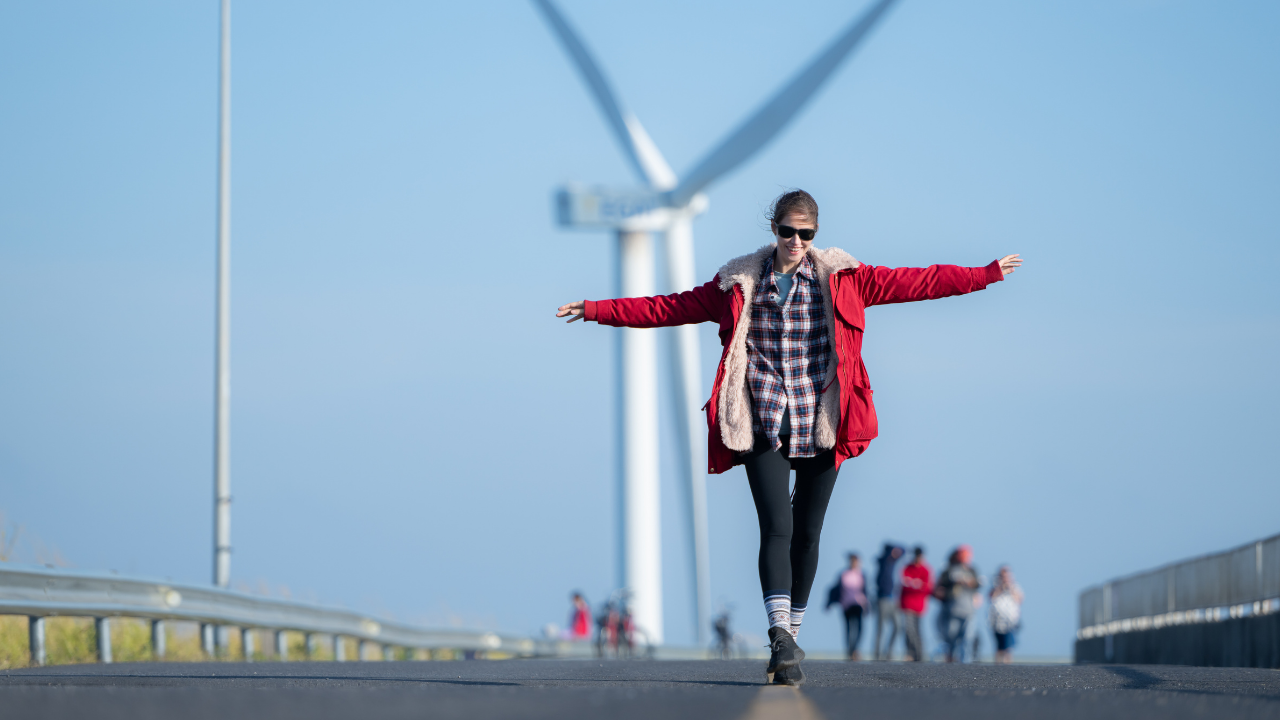 A young woman balances joyfully on a rural road with a wind turbine in the background, subtly illustrating public awareness of how wind energy works.