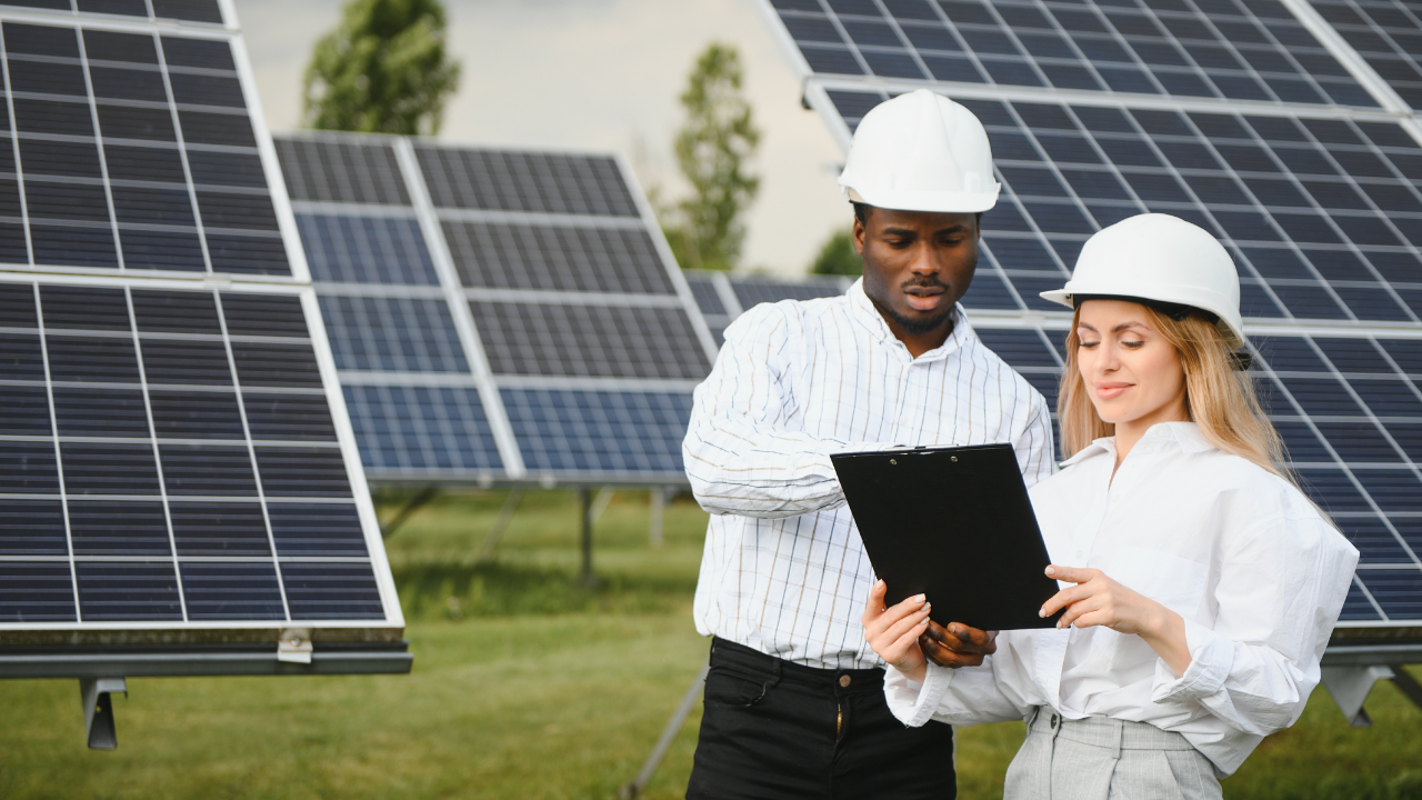 Two engineers, one pointing at a clipboard and the other reviewing it, stand in front of large solar panels. The phrase "solar panels" reflects their focus on data review and collaboration in renewable energy projects.