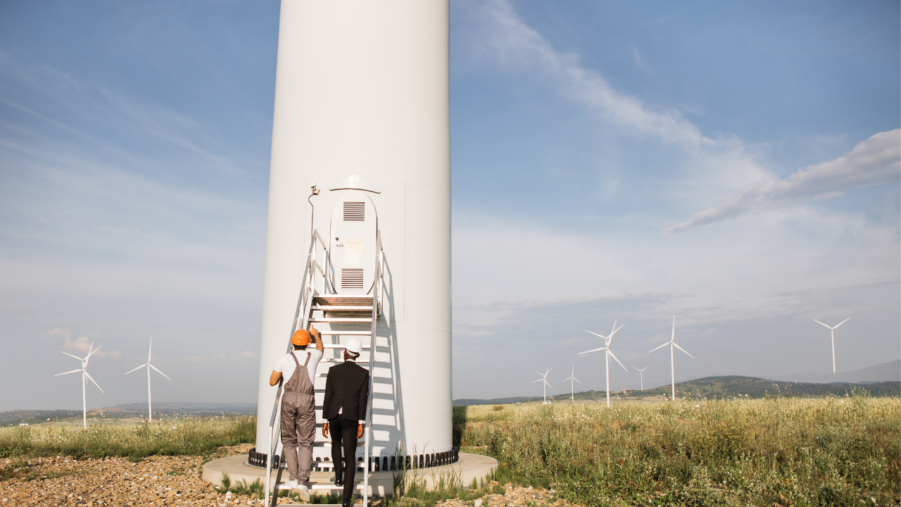 A worker and a suited engineer inspect the base of a turbine, symbolizing technical engagement in understanding how wind energy works.