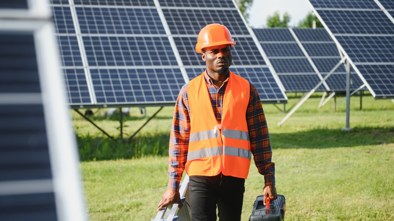A technician in an orange vest and helmet carries tools across a solar field. "Solar panels" underscores the active, hands-on maintenance work critical to the success of green infrastructure.