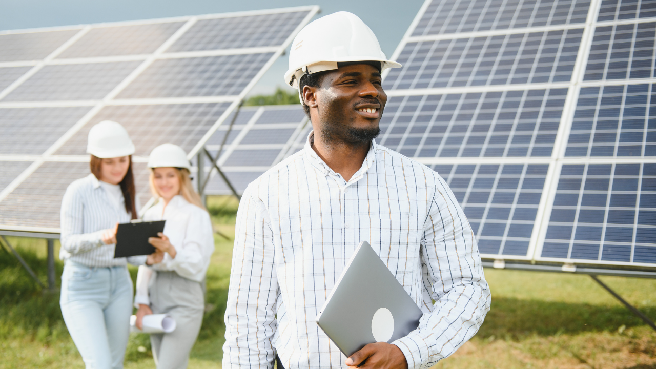 Un ingeniero sonriente con una laptop se destaca en primer plano, mientras dos colegas en segundo plano trabajan juntas entre paneles solares. "Paneles solares" simboliza un entorno laboral positivo y orientado al desarrollo sustentable.