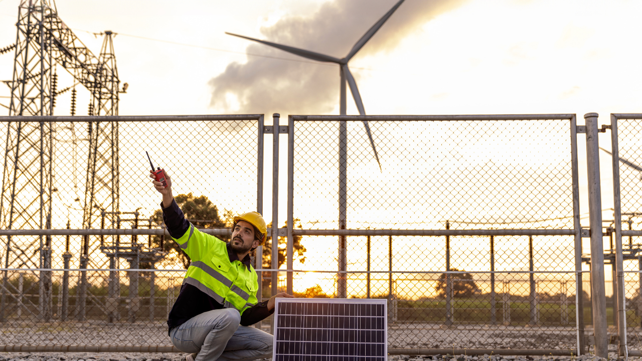 Un técnico agachado sostiene un walkie-talkie junto a un panel solar bifacial, con turbinas eólicas al fondo. La frase "paneles solares bifaciales" refleja la integración de distintas fuentes de energía limpia en la infraestructura nacional.