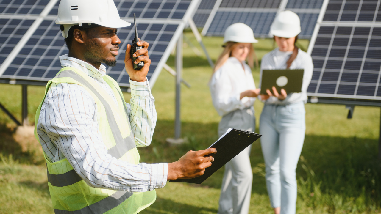 Un operario con chaleco reflectante utiliza un walkie-talkie mientras consulta una planilla, con paneles solares y colegas de fondo. La frase "orientación e inclinación" transmite el enfoque detallado y organizado del trabajo en campo.