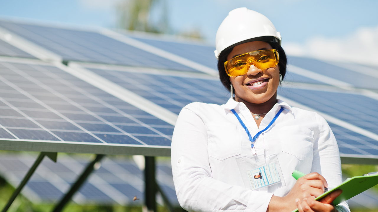 Una mujer sonriente con casco y gafas protectoras sostiene una carpeta verde frente a los paneles solares, proyectando confianza y conocimiento sobre la eficiencia de tus paneles solares.