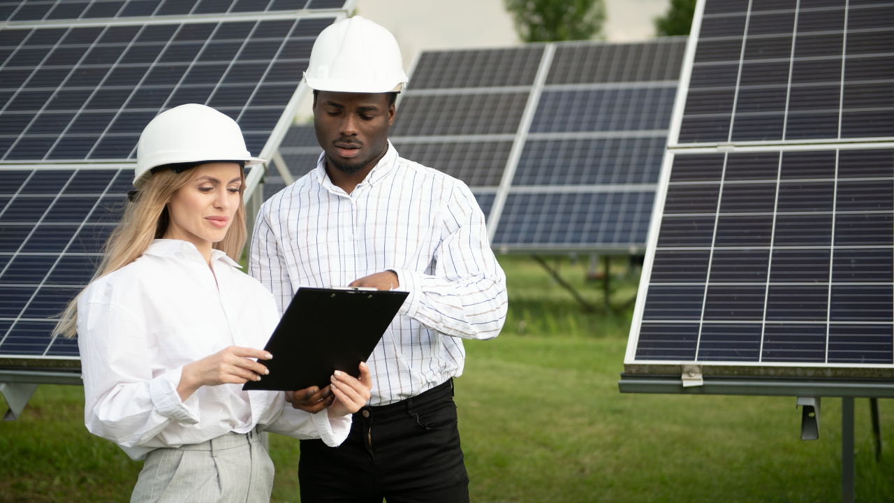 Two engineers, one male and one female, stand in front of solar panels reviewing data on a clipboard, symbolizing collaboration and progress in solar energy applications.