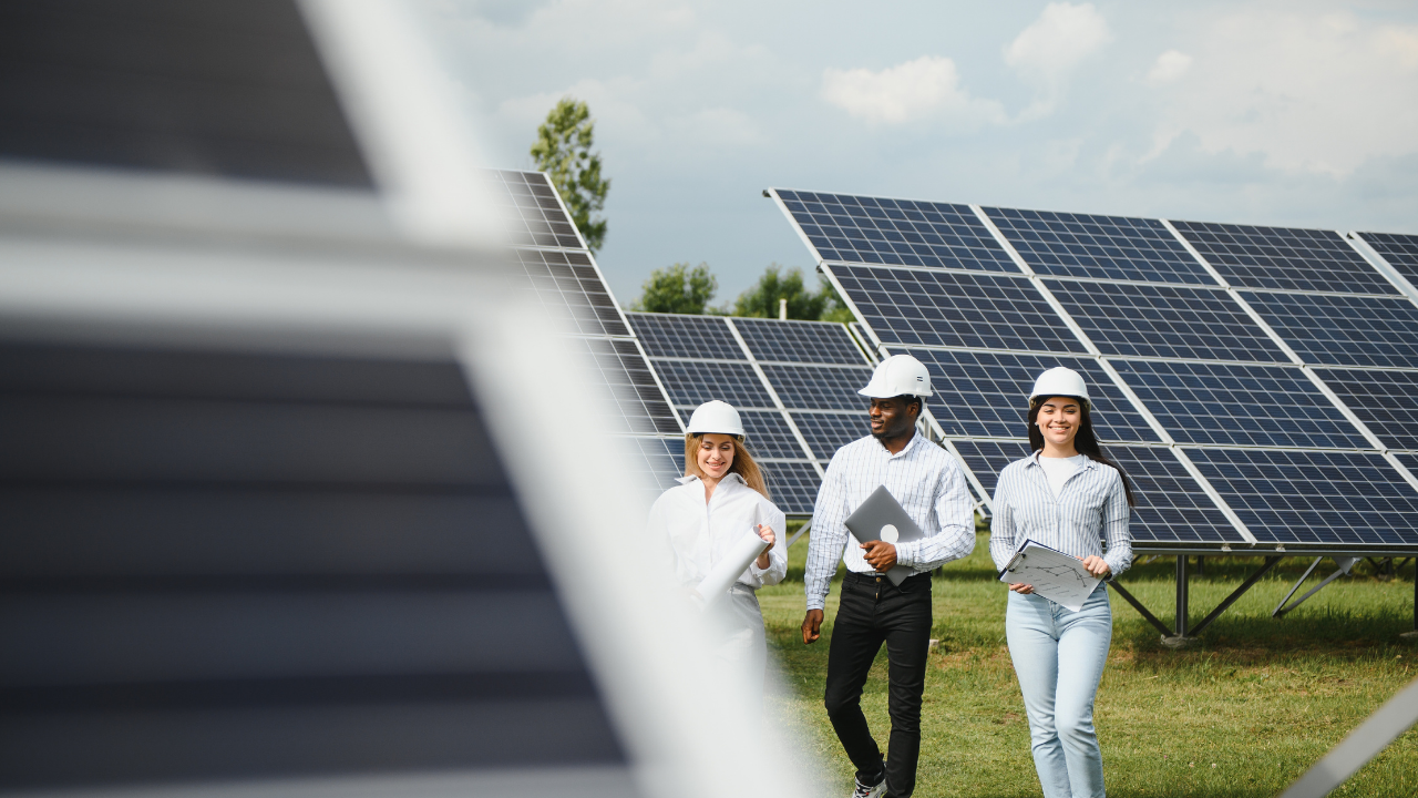 Un equipo mixto camina entre paneles solares, portando tabletas y documentos, destacando el enfoque dinámico y juvenil hacia la energía solar en el contexto español.