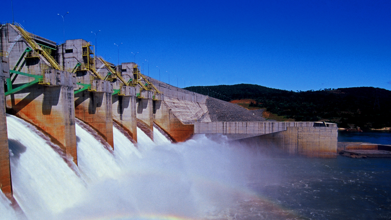 Water gushes forcefully through the dam gates under a clear blue sky, highlighting the dynamic energy production of hydroelectric power plants.